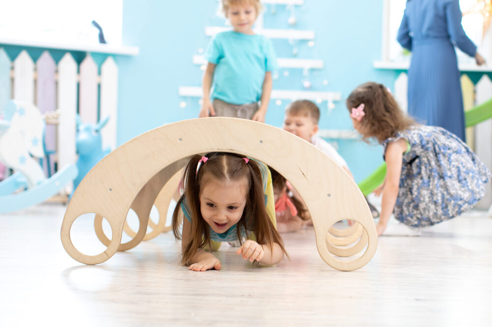 A smiling young girl crawling under a wooden play arch with friends in a bright playroom.