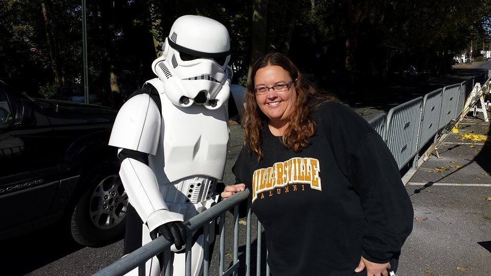 A woman in a Millersville Alumni sweatshirt posing with a Star Wars Stormtrooper outdoors.