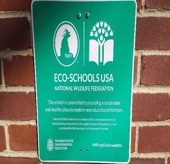 A green Eco-Schools USA sign mounted on a red brick wall, declaring the school's commitment to sustainability and environmental education