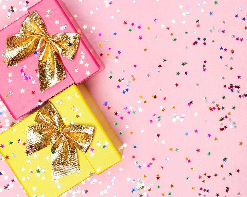 Pink and yellow gift boxes with gold bows surrounded by star confetti on a pink background.