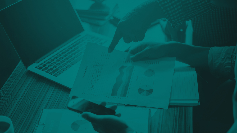 Teal‑tinted image of two people reviewing printed charts and graphs at a desk with a laptop, pointing to data on a report.