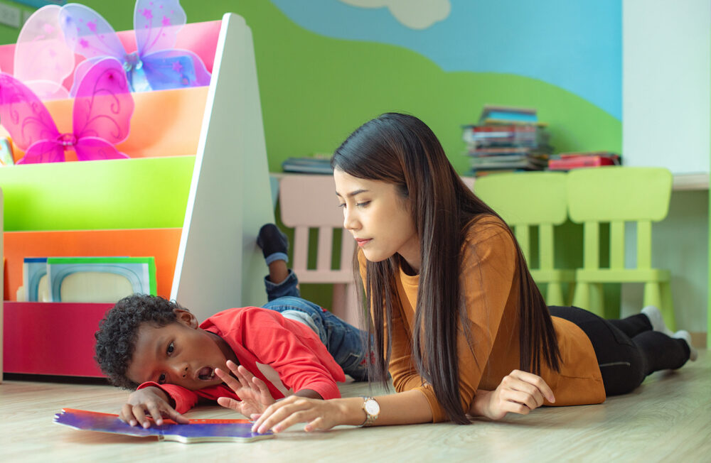 dark haired woman reading to upset child wearing red shirt and making frustrated face. they are in a green playroom with fairy wings and green chairs, and laying on the floor