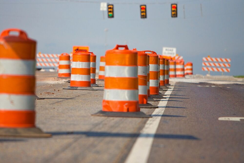 A row of orange and white striped construction barrels blocking lanes on a paved road ahead of an intersection with traffic lights
