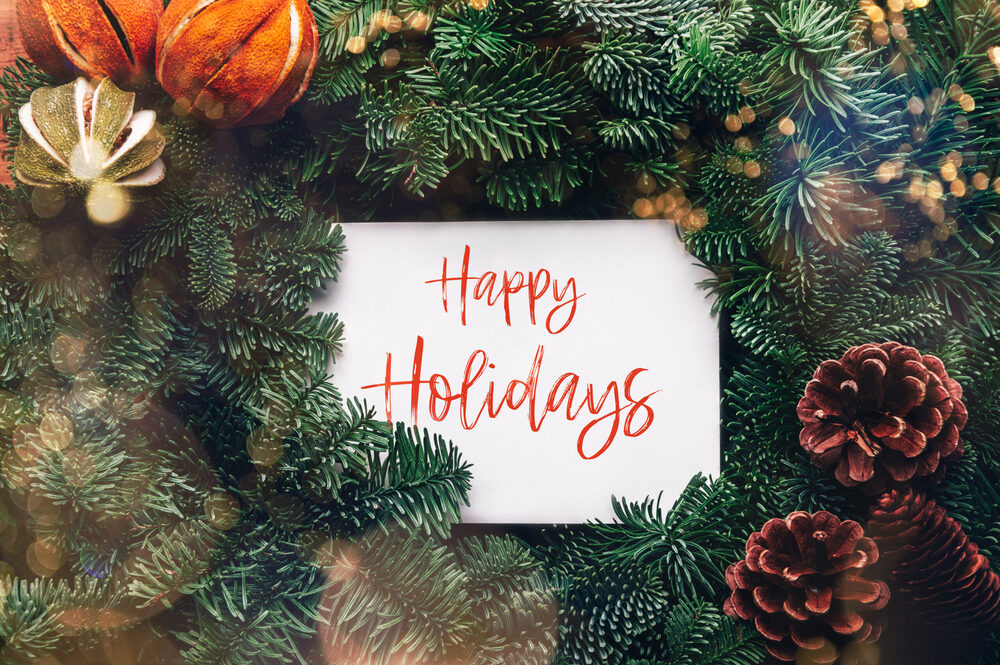 "Happy Holidays" Text in red cursive font on a background of a green wreath with pinecones and ornaments