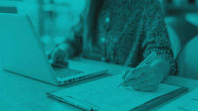 A person writes on a document with a pen while working on a laptop, styled with a monochromatic teal overlay.