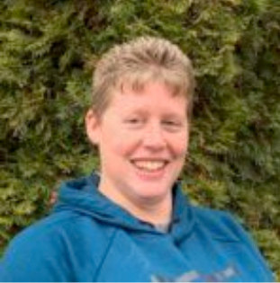 blonde person with short hair in a blue hoodie in front of green bushes