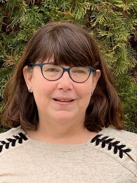 brown haired woman with glasses in a tan sweater in front of green bushes