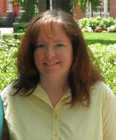 woman with brown hair and bangs smiles in front of green bushes, grass, and trees while wearing a light yellow button up shirt