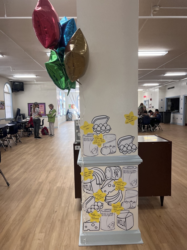 picture of a column in the VSDB cafeteria with cutout images of various food items taped to it. there are several yellow stars taped in this area also, each with writing. stars read: "each food item equals 100 meals," "great job!" , "every team has food in their donation box. well done!", "green team is winning in cash donations," and other positive messages.