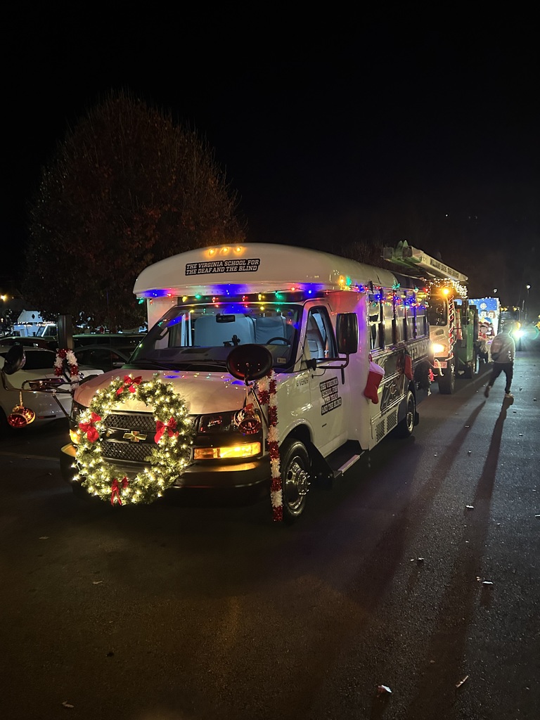 A side view of a decorated white bus at night, also bearing the text "The Virginia School for the Deaf and the Blind." The bus is adorned with colorful holiday lights, a wreath on the front grille, and candy cane decorations on its mirrors. Other decorated vehicles are visible in the background.