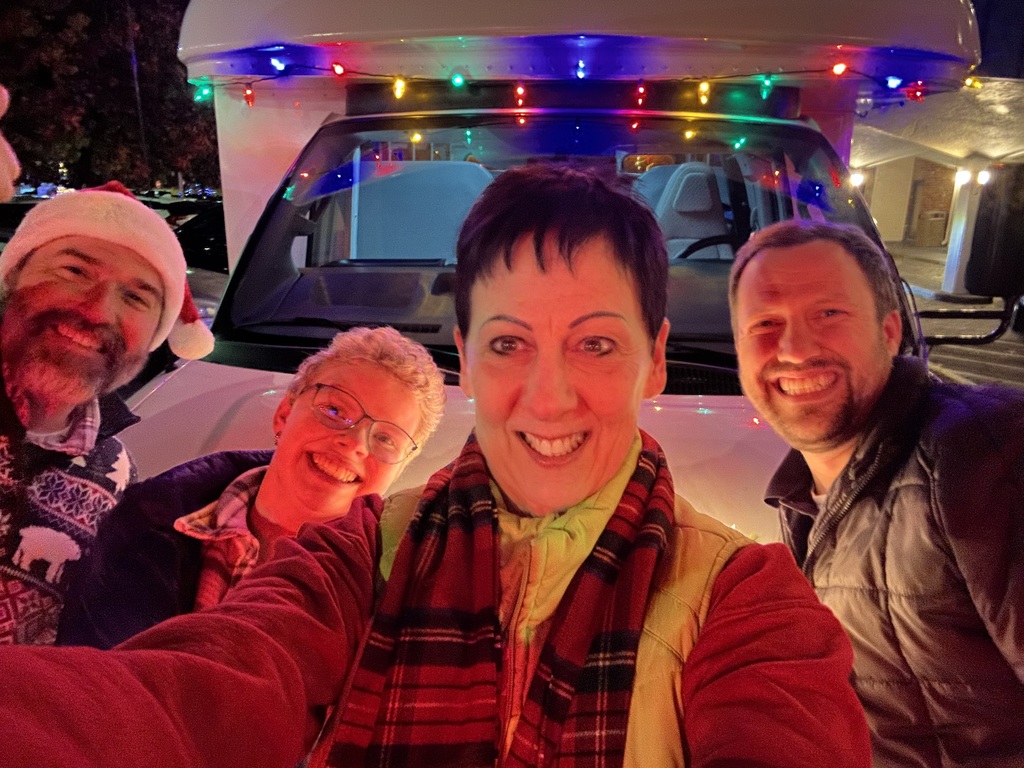The image features a group of four people posing for a selfie in front of a white vehicle, which is adorned with colorful string lights. The picture is taken at night, and the festive lights create a warm and cheerful ambience. The individuals appear happy, smiling at the camera. The person on the far left is wearing a Santa hat, adding to the holiday theme. Trees and lights can be faintly seen in the dark background, suggesting an outdoor setting, and a building with lights is visible on the far right
