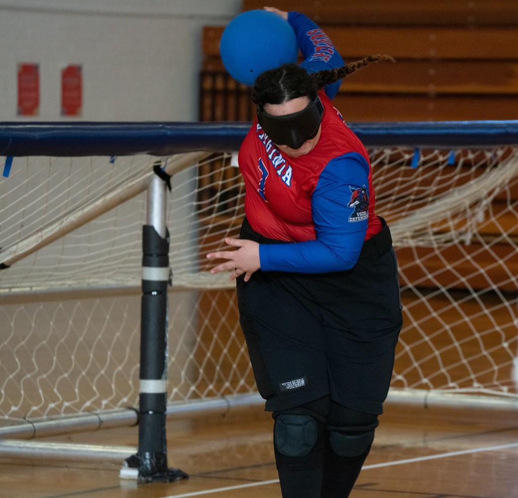 The image shows a person participating in a goalball game, a sport designed for athletes with visual impairments. The athlete is captured in motion, wearing a black blindfold and protective gear including a red jersey with blue long sleeves and black knee pads. The individual is in the process of throwing a blue ball with one hand, showcasing an athletic posture. Behind the player is a goal with a net and white netting spread throughout the frame. The setting is an indoor gymnasium with wooden floors and brown bleachers in the background.
