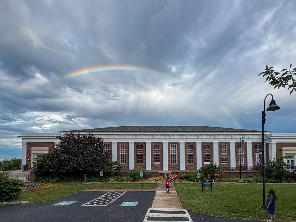 Coach Patti with the girls volleyball team doing running drills outside the gym, with a rainbow in the back ground. On the left hand side you can see the barest glint of a second rainbow. 
