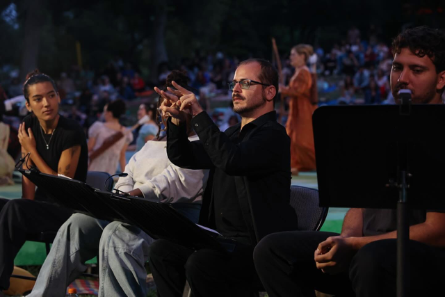 Neil is signing in front of stage and wearing a black button down shirt. A play is on stage in the background.