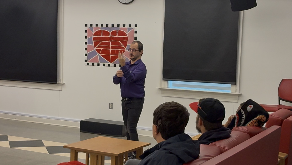Neil is signing in front a room while students watch from a couch on the right.