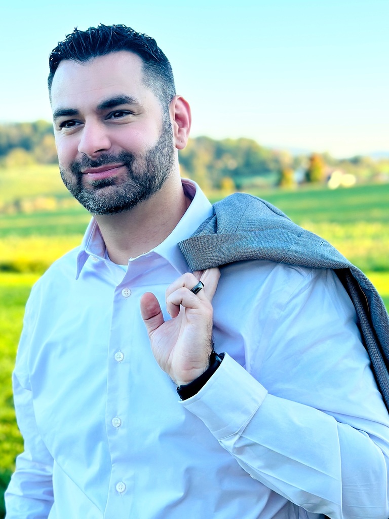 Michael is standing outside and smiling with a blazer slung over his shoulder.