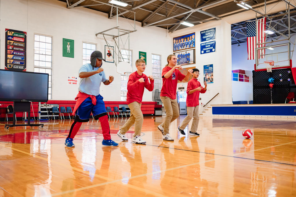: Four students perform a choreographed dance on the gym floor during the pep rally. They are in motion, showing energy and team spirit. Banners and an American flag hang in the background.