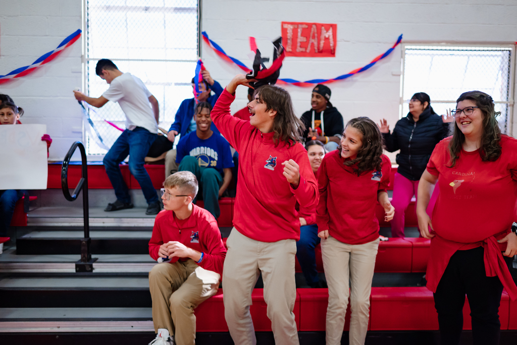 A group of students and staff cheer enthusiastically from the bleachers, some standing and waving, others holding signs. One student lifts a hat in celebration, and red and blue streamers decorate the walls.