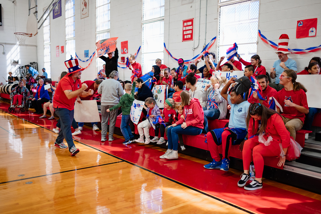 A group of students and staff cheer enthusiastically from the bleachers, some standing and waving, others holding signs. One student lifts a hat in celebration, and red and blue streamers decorate the walls.