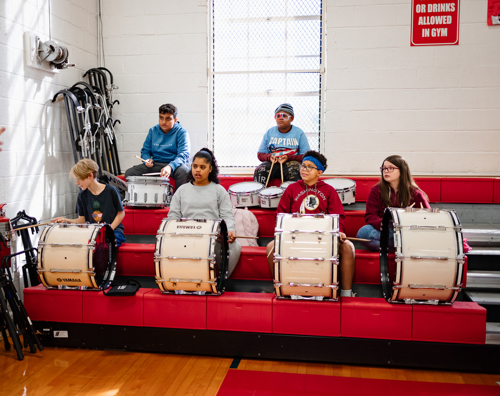A student drumline sits in the bleachers, each with a drum in front of them. They are focused and ready to play, contributing to the pep rally’s lively atmosphere.