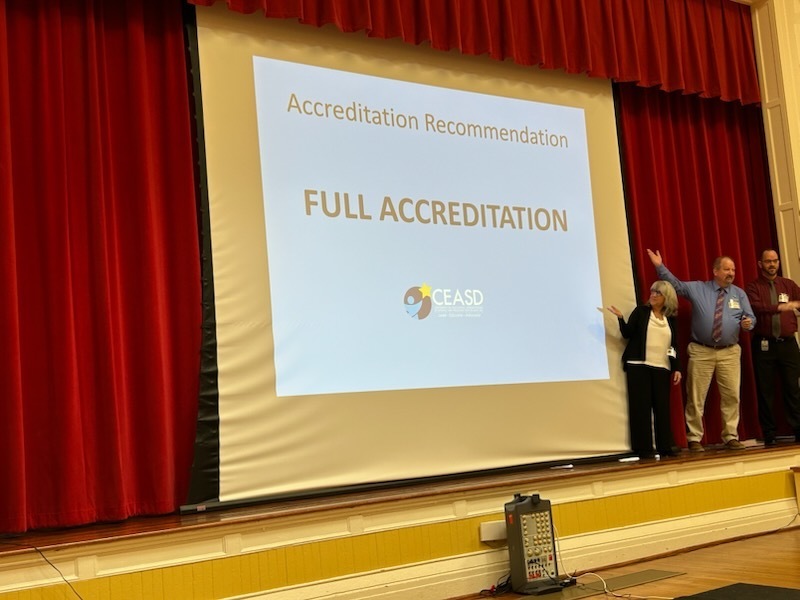 Presenters stand on a stage in front a screen that reads  "Accreditation Recommendation Full Accreditation"