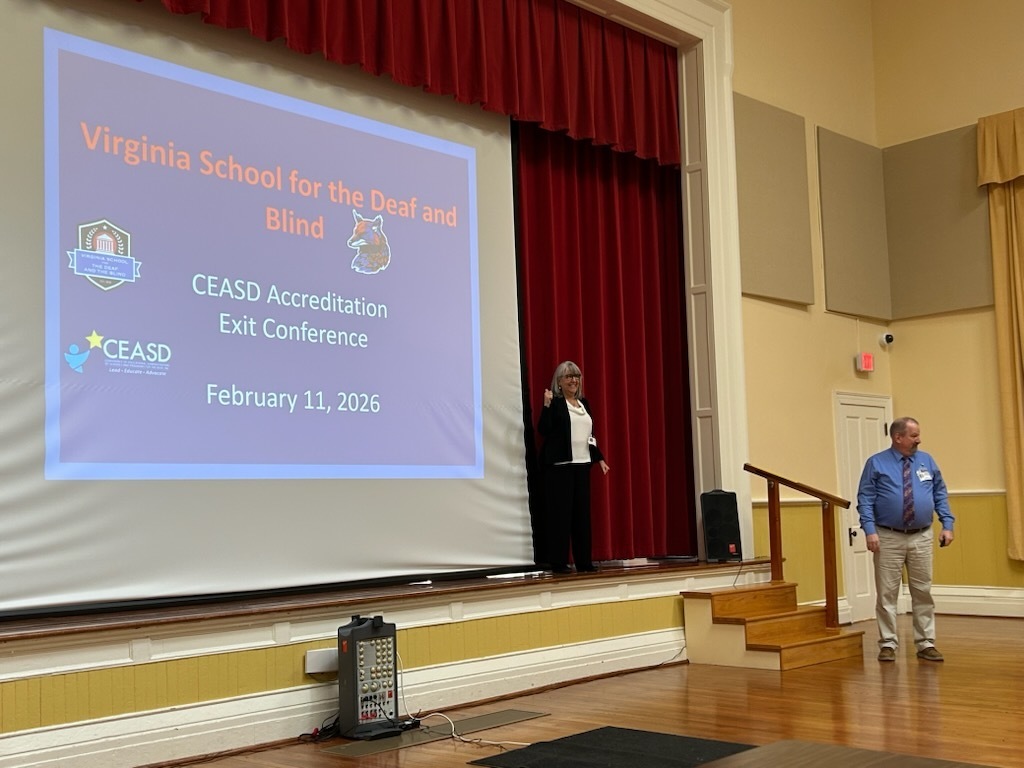 Presenters stand on a stage, and  in front a screen that reads "Virginia School for the Deaf and the Blind CEASD Accreditation Exit conference  February 11, 2026"