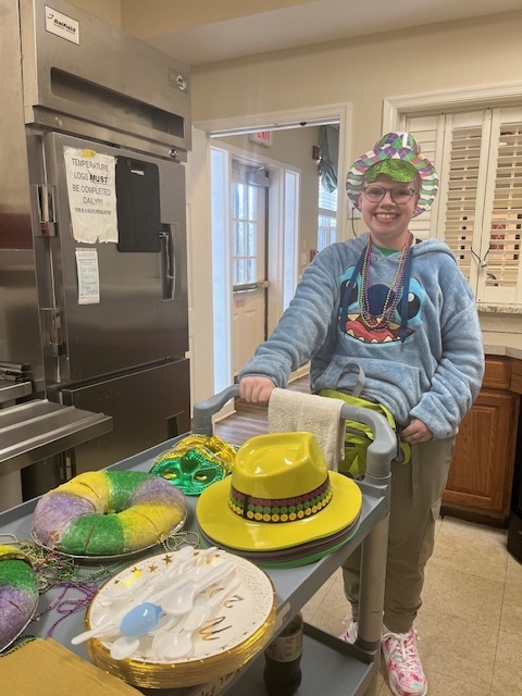 Brianna is standing in a stitch hoodie, a Mardi Gras themed hat, and has beads around her neck. She is pushing a cart filled with Mardi Gras favors.
