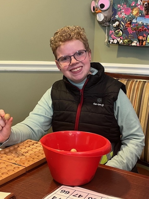 Brianna is sitting a table and smiling at the camera. There is a red bucket filled with small balls for a game.