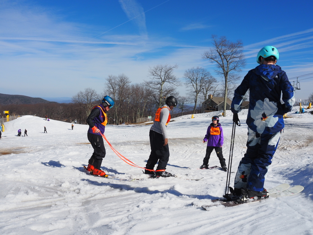 A blind skier is attached by ropes at the legs to a guide as they ski down a slope. A ski guide is also in front of them.