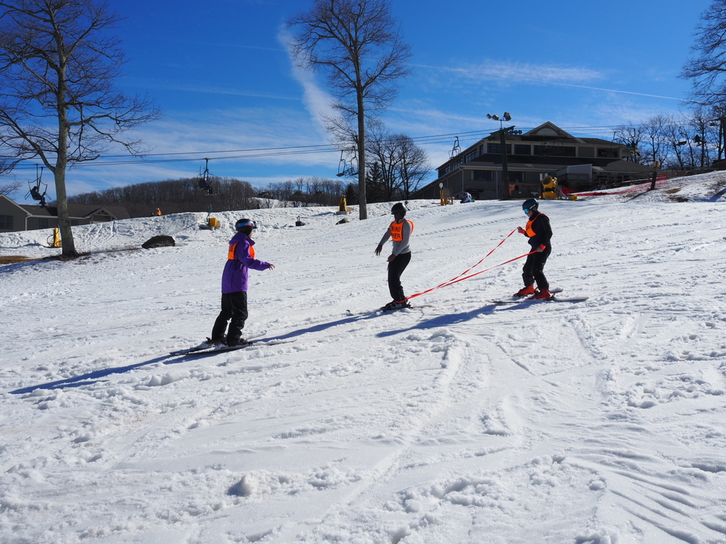 A blind skier is attached by ropes at the legs to a guide as they ski down a slope . A second guide is in front.