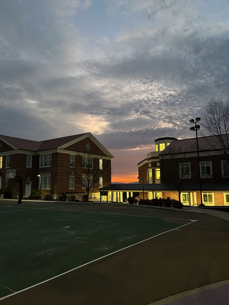 A bright orange sunrise begins in the distance. The sky is cloudy and tinged pink near the orange. The VSDB buildings are in the forefront with the lights from the building shining bight.