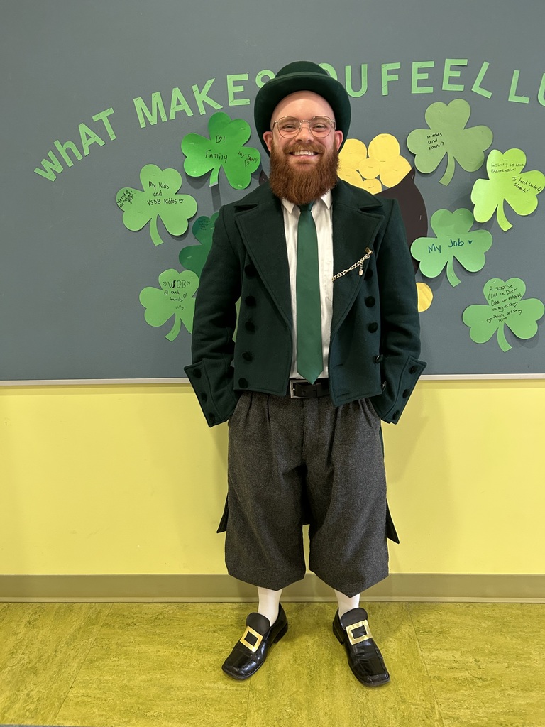 A Staff member with a long orange beard  smiles in front of a board that has paper 4 leaf clovers on it and says "What makes you feel lucky".  The man is in a green tophat,  green coat, wearing a green tie, and has folden shoe buckles on his black loafers - it is a beautiful leprechaun costume. 