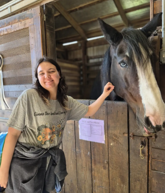 A student is smiling next to a large horse that is looking our of their stall.
