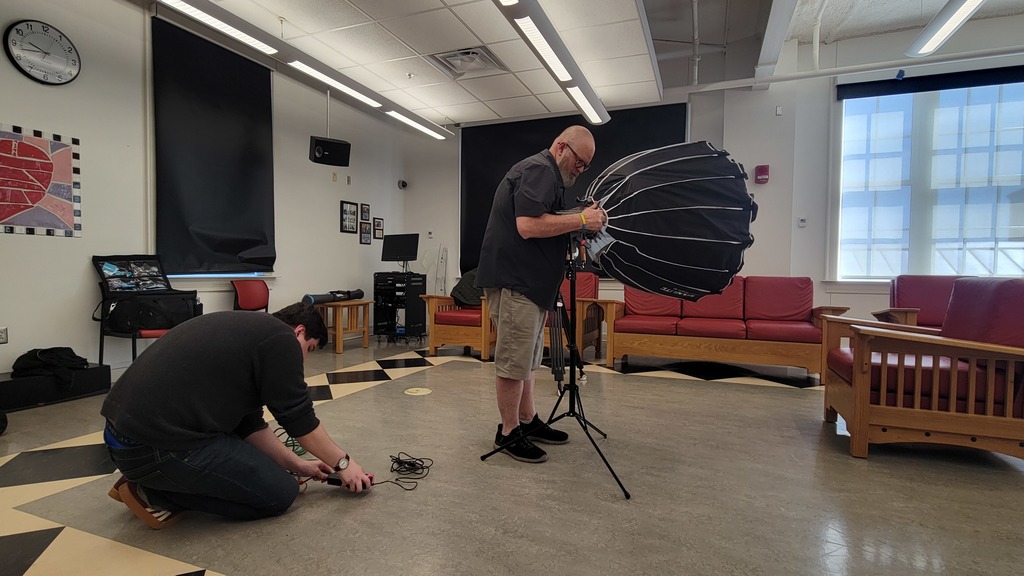 Peeople are setting up filming equipment - one person is plugging in a charger while the other sets up a lighting rig.