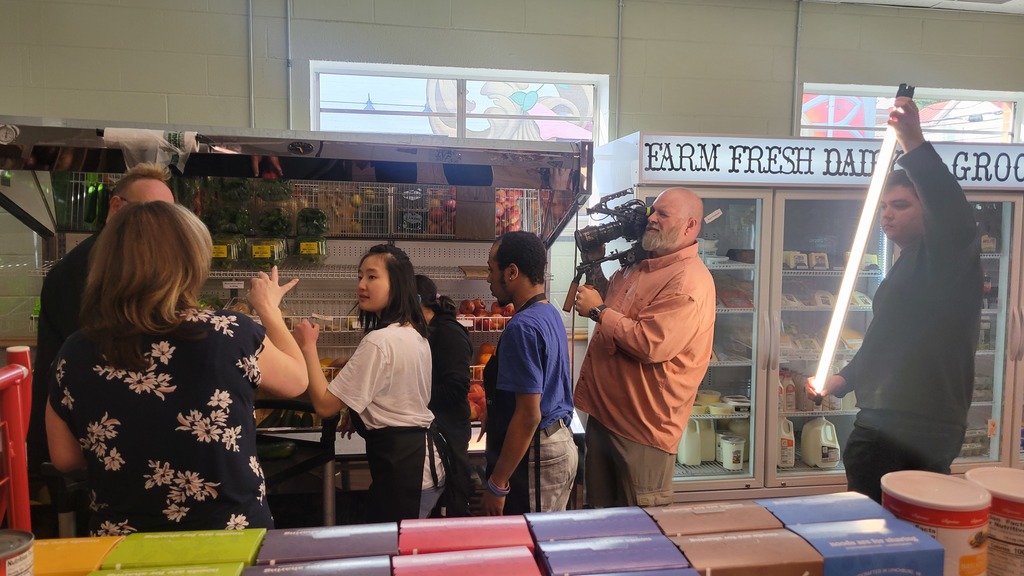 Students and staff are gathered in a grocery store and are signing to each other. There is a camera man recording everything, and a man holding a light stick tpwards to group.