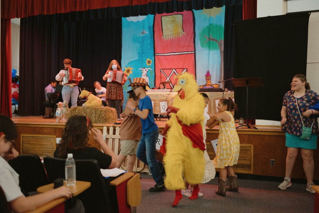 Students are performing on stage, while others are dancing at the base of the stage with someone in a full chicken costume.