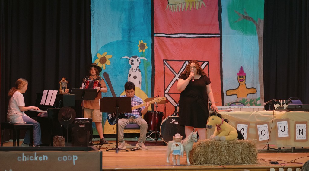 Students performing on a stage with country decor in the background. There is a singer, a guitar player, a piano player, and an accordian player.