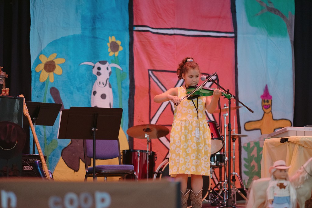 A young student is on stage playing a green fiddle. Country decor is in the background.