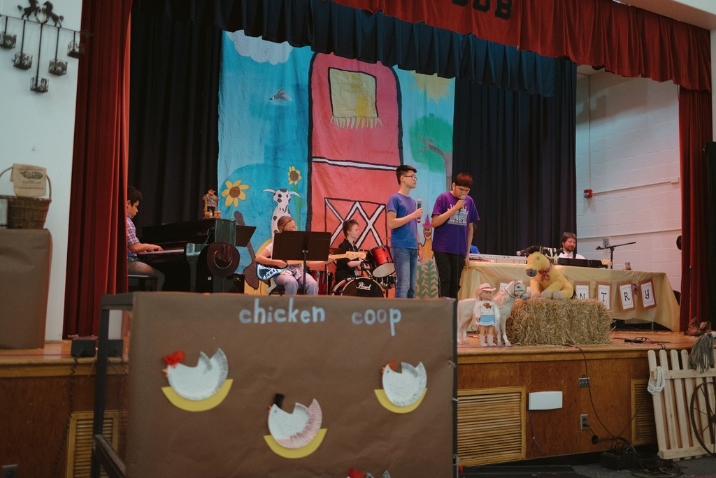 students are performing on stage. In the foreground we can see a "chicken coop" in the backrgound there is country decor.