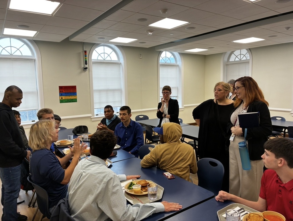 male high school students eating lunch in the cafeteria and signing to Dr. Maxlow. Dr. Maxlow is smiling as they talk to her. 