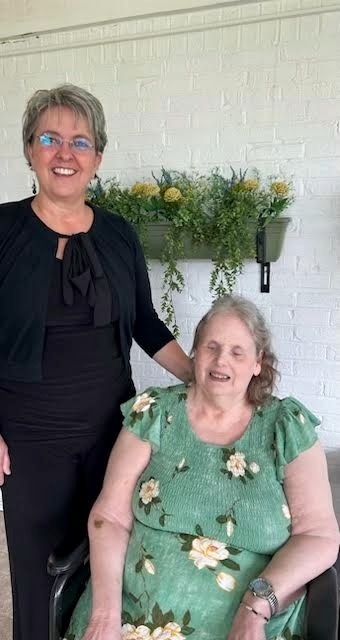 Superintendent Charity Warigon stands beside Faith Cummings, who is seated, both smiling toward the camera. They are in front of a white brick wall with a wall‑mounted planter filled with green foliage and yellow flowers. Charity wears a black outfit and glasses; Ms. Cummings wears a green floral dress. The setting appears to be an indoor or covered patio space.