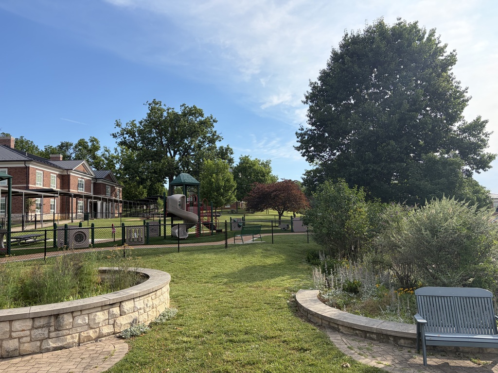 A photo taken outside of a playground area. The sky is a bright blue, the trees are full and green, there are lavender plants and flowers in a bricked off area, and there is one red tree in the center of the photo. There is a bench in the foreground, and a brick building in the background.