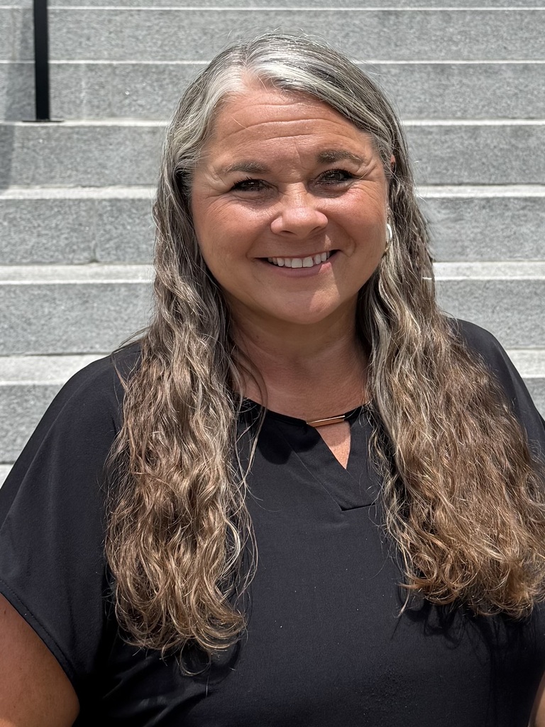 A woman with long, grey hair smiles in front of stone steps. She is wearing a black shirt.