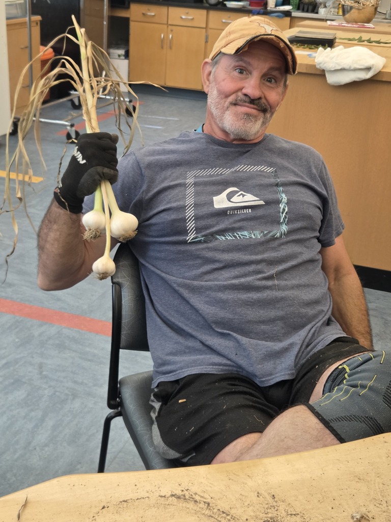 Todd is holding up three cloves of garlic, hes inside and wearing a hat and T-shirt.