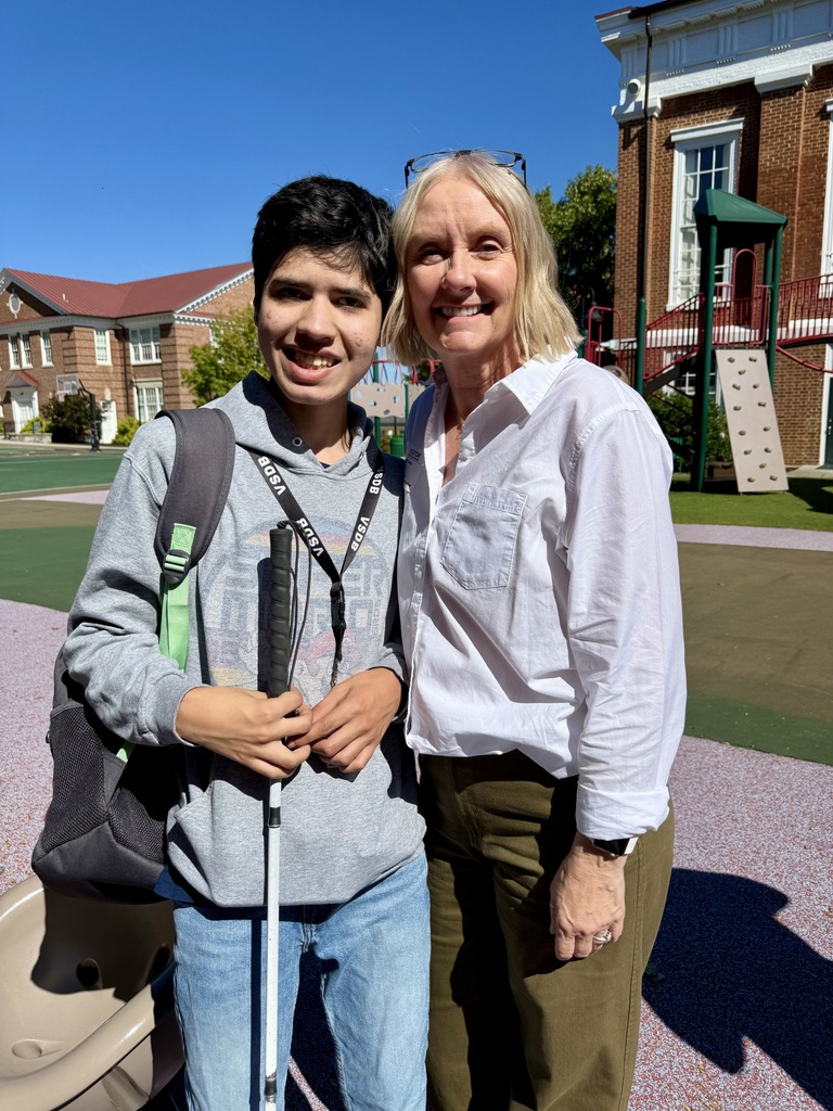 Kerry with teenage student holding his cane. they are outside near the playground.