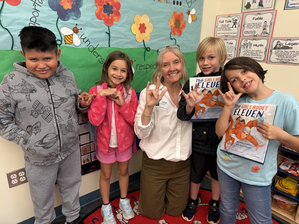 four elementary students with Kerry in a classroom. several students are doing the I love you sign and teo are holding copied of the Gallaudet Eleven