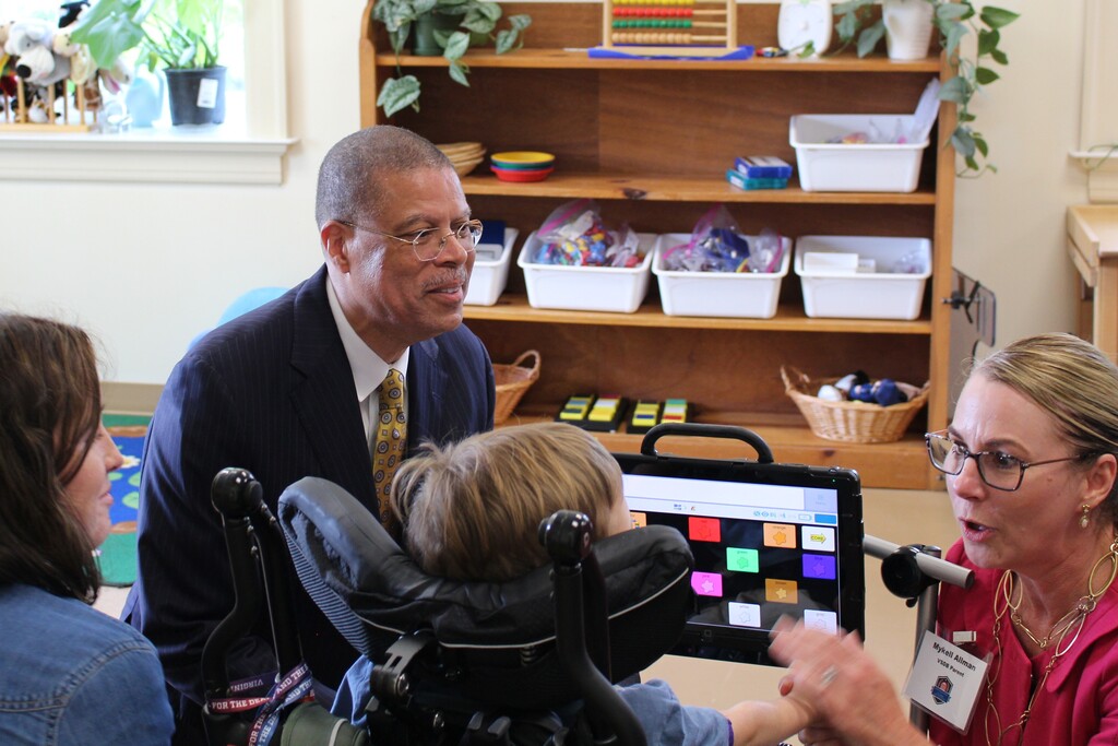 Dr. Jeffrey O. Smith is visiting a classroom and is smiling towards a woman in pink who ius interacting with a young student in wheelchair. A woman is smiling off to the left.