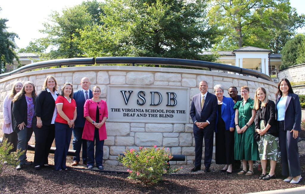 A group is standing outside in front of the VSDB Sign that reads "VSDB The Virginia School for the Deaf and the Blind".. From left to right we have: Mary Eppard, Jessica McMahon, Julie Conner, Tanya  Johnson, Scott Alleman, Mykell Alleman, Dr. Jeffrey O. Smith, Chairty Reedy Warigon, Dr. Selmo Warigon, Lindsay Burris, Abby Edwards, and   Andrea Mariscal-Guzman