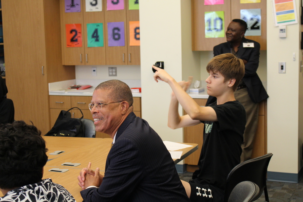 Dr. Jeffrey O. Smith is sitting between students in class smiling. One student is signing a response. A man stands smiling in the background. 
