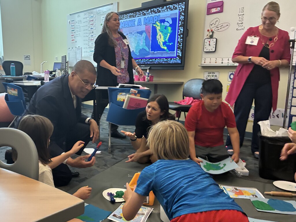 Dr. Jeffrey O. Smith is in a classroom and is handing a tool to a young student to help her make a Play Dough Map. multiple students are working on their maps, and multiple adults are standing in the background. 